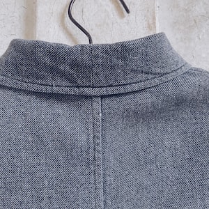 May include: A close-up of the back of a dark gray denim jacket with a collar. The jacket has a seam down the back and a single stitch along the collar.