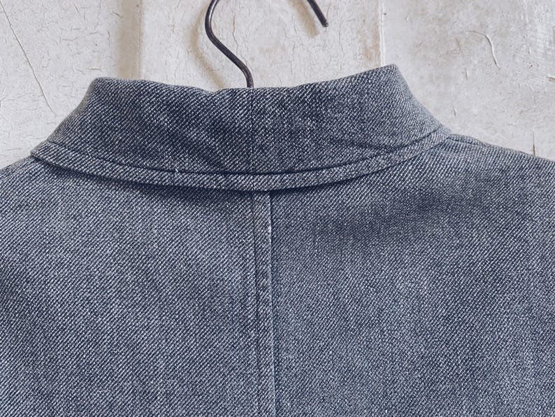 May include: A close-up of the back of a dark gray denim jacket with a collar. The jacket has a seam down the back and a single stitch along the collar.