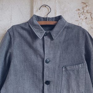 May include: A gray denim work jacket with a button-up front and a single patch pocket on the left side.