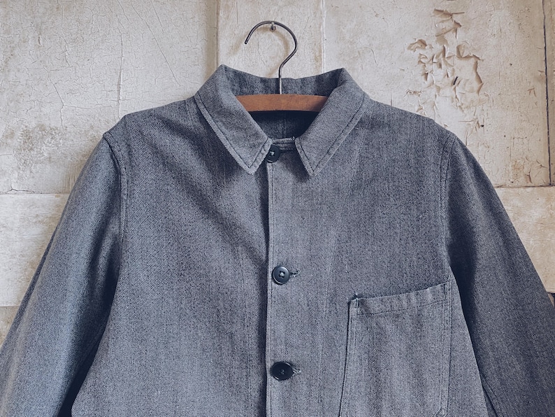 May include: A gray denim work jacket with a button-up front and a single patch pocket on the left side.
