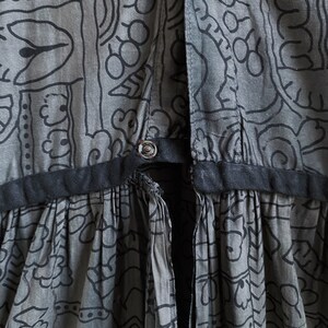 May include: A gray cotton dress with a black waistband and a black and white geometric print. The dress has a tie at the back.