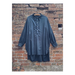 May include: A dark blue long-sleeved shirt with a button-down collar and a loose fit. The shirt is displayed on a wooden hanger against a weathered brick wall. The shirt has a classic, minimalist design.