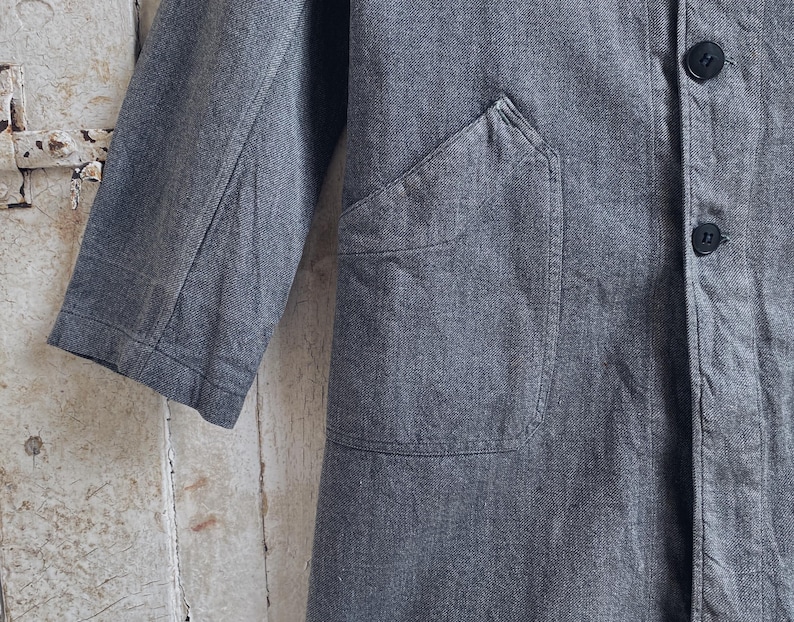 May include: A gray denim jacket with a large patch pocket on the front. The jacket has a button closure and two buttoned pockets on the front.