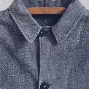 May include: A gray denim jacket with a collared neckline and two black buttons. The jacket is hanging on a wooden hanger.