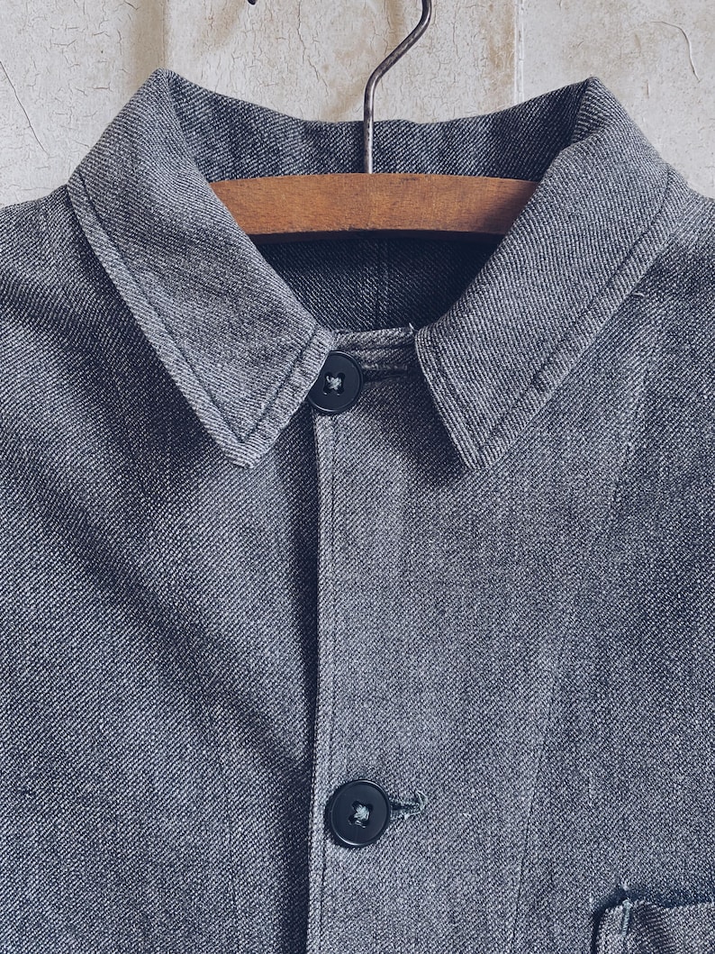 May include: A gray denim jacket with a collared neckline and two black buttons. The jacket is hanging on a wooden hanger.