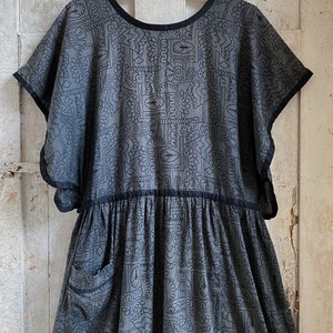May include: A gray cotton dress with a black geometric pattern and black trim. The dress has a round neckline, short sleeves, and a gathered skirt. It features a single pocket on the left side.