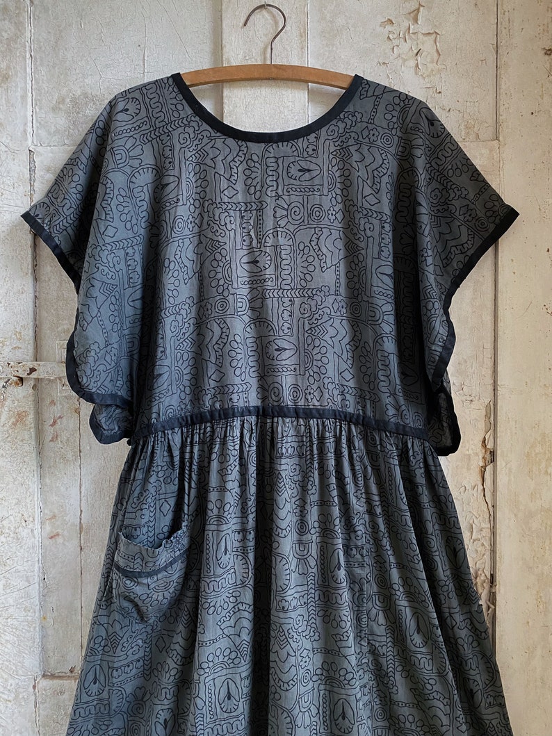 May include: A gray cotton dress with a black geometric pattern and black trim. The dress has a round neckline, short sleeves, and a gathered skirt. It features a single pocket on the left side.