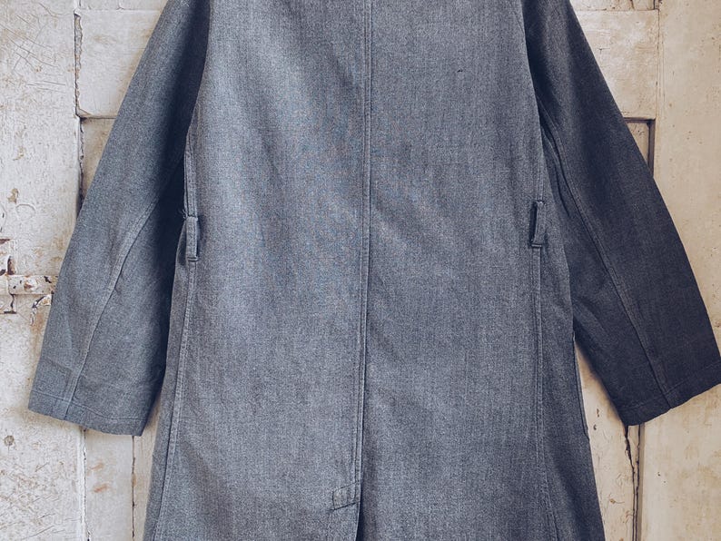 May include: A long, gray, denim jacket with a single-breasted closure and two pockets on the front. The jacket has a slightly textured surface and a relaxed fit.