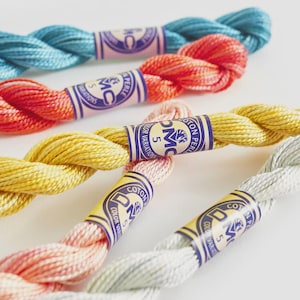May include: Close-up of colorful DMC pearl cotton embroidery floss skeins. The skeins are in shades of blue, orange, yellow, pink, and gray. Each skein has a paper label with the DMC logo and text. The floss is tightly twisted.
