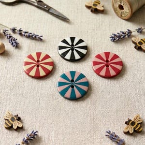 May include: Four circular buttons featuring a two-hole center and a radial striped pattern in black and white, red and cream, red and pink, and teal and pink. The buttons are arranged in a cluster alongside small wooden bee-shaped embellishments.