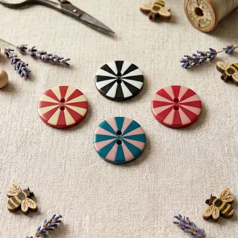 May include: Four circular buttons featuring a two-hole center and a radial striped pattern in black and white, red and cream, red and pink, and teal and pink. The buttons are arranged in a cluster alongside small wooden bee-shaped embellishments.