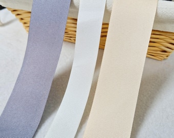 May include: Three different colors of ribbon, gray, white, and beige, are displayed in a wicker basket. The ribbons are all the same width and have a ribbed texture.