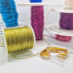May include: Spools of metallic thread in gold, purple, pink, and blue. The spools are on white plastic bobbins. There is a small bag of gold thread.
