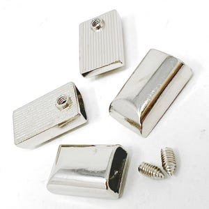 May include: A collection of silver-toned metal furniture feet and screws. Four rectangular feet with rounded edges and threaded inserts are displayed, two with a ribbed texture. Two small screws are also visible.
