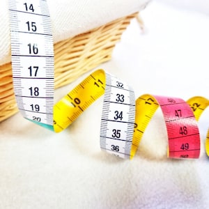 May include: A close-up of three measuring tapes. The first tape is yellow and white, the second is white and black, and the third is pink and white. The tapes are coiled and overlapping.
