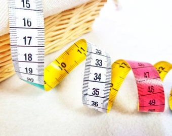 May include: A close-up of three measuring tapes. The first tape is yellow and white, the second is white and black, and the third is pink and white. The tapes are coiled and overlapping.