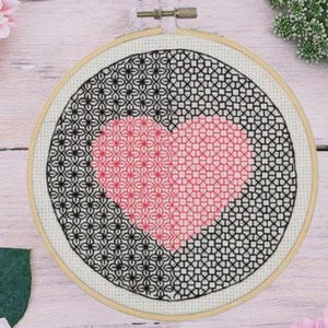 May include: A cross-stitch embroidery hoop featuring a pink heart design in the center, surrounded by a black geometric pattern. The embroidery is set in a wooden hoop. The background includes flowers and greenery.