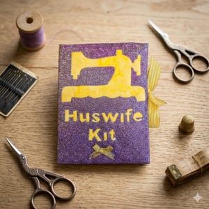 May include: A purple glittery Huswife Kit with a yellow sewing machine design and text. The kit is surrounded by sewing tools, including scissors, needles, a thimble, and a measuring tape, all arranged on a wooden surface.