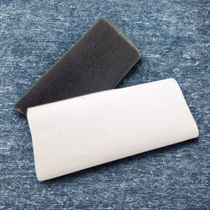 May include: Two rectangular erasers, one dark gray and one white, are displayed against a blue textured background. The dark gray eraser is angled, partially overlapping the white eraser. The erasers appear to be new.