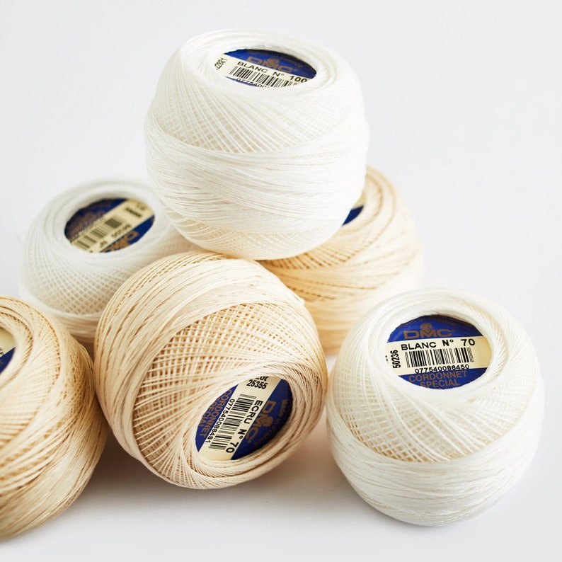 May include: Close-up of six spools of DMC embroidery floss. The spools are white and beige. The labels on the spools read "Blanc N&deg; 100" and "Ecru N&deg; 70".