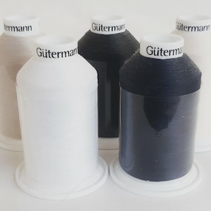 Op de afbeelding: Vijf klossen Gütermann naaigaren gerangschikt in een groep. De klossen hebben witte kunststof voetjes en bovenkanten met de merknaam in het zwart gedrukt. De kleuren garen zijn wit, zwart, donkerblauw, beige en gebroken wit, elk omwikkeld.