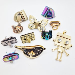 May include: Assortment of colourful metal bag clasps in various shapes, including a heart, butterfly, cat, robot, bird, and woven design. The clasps are made of gold-toned metal with some featuring iridescent finishes.