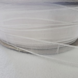 May include: A roll of white sheer ribbon. The ribbon is tightly wound, with visible horizontal lines. The ribbon has a fine, open weave, creating a delicate, translucent appearance. The ribbon is likely used for crafts, gift wrapping, or decorative purposes.