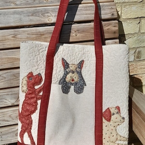 May include: Cream quilted tote bag with red handles and trim. The bag features appliqued dog designs in red, black, and tan. The bag is made of fabric and has a rectangular shape.