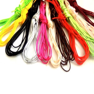 Può includere: Una collezione di cordini colorati in varie tonalità, tra cui verde, giallo, nero, bianco, rosa, marrone, rosso e crema. Ogni cordino è annodato in alto, con le estremità sciolte che si estendono verso il basso. I cordini sono disposti su uno sfondo bianco.