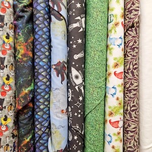 May include: A selection of rolled fabric bolts featuring diverse patterns. Designs include a clown print, a galaxy-like pattern, blue scales, a wizard, a rocket ship with the text "Adventure Beyond", grass, gnomes, and a floral pattern. The fabrics are in shades of grey, blue, green, and purple.