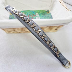 May include: A grey leather strap with a gold chain detail. The strap has a metal ring at the end.