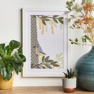 May include: A framed cross-stitch artwork featuring a bird perched on a tree branch. The artwork has a white frame and is displayed on a wooden surface. Decorative plants and a vase are also present.