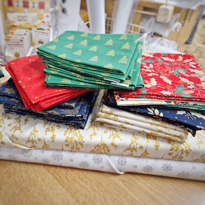 May include: Stacks of Christmas-themed fabric in various colors and patterns. Green fabric with gold Christmas trees, red fabric with gold dots, and blue fabric with gold floral designs are visible. Other fabrics include white with gold floral and white with silver snowflakes.