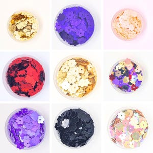 May include: Assortment of nine containers filled with colourful flower-shaped sequins. The sequins are in various colours, including gold, purple, red, black, and pink.