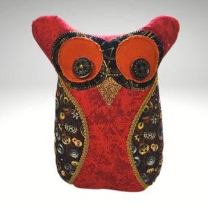 May include: A decorative owl ornament. The owl is predominantly red, with orange eyes and black and gold detailing. It has button eyes and a body patterned with gears and gold trim. The owl's wings are red.