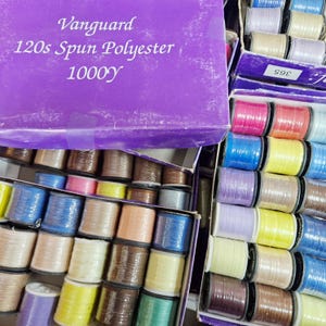 May include: A purple box with the text "Vanguard 120s Spun Polyester 1000y" on the top. The box is filled with spools of thread in various colours, including red, blue, green, yellow, and brown.