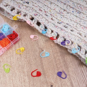 Puede incluir: Un organizador de plástico transparente lleno de marcadores de punto de colores, incluyendo rosa, verde, naranja y azul. También se ve una sección de un artículo de ganchillo moteado, blanquecino, con marcadores de punto.