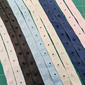 May include: A selection of fabric snap tape strips in diverse colours, such as white, black, light blue, and navy. Each strip has evenly spaced snap closures, suitable for sewing. The strips are arranged diagonally across a green cutting mat.