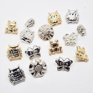 May include: A collection of decorative metal embellishments in gold and silver tones. The assortment includes owl, frog, butterfly, skull, and flower designs. These small, detailed pieces are suitable for various craft projects.