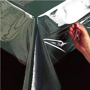 May include: Clear plastic tablecloth with a smooth, shiny surface. The tablecloth is draped over a table and a hand is holding a corner of the tablecloth.