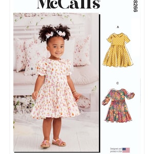 May include: A sewing pattern for a girl's dress with three different views. View A is a yellow dress with white polka dots, View B is a white dress with pink and red floral print, and View C is a multi-coloured floral print dress. The pattern is designed and printed in the USA. McCall's M8266.