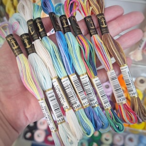 May include: A collection of eight Anchor brand embroidery floss skeins in various variegated color combinations including pastel pink, yellow, blue, green, and brown, each featuring a black paper label with an anchor logo and a barcode.