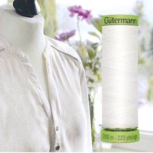 May include: A white Gütermann thread spool with a green base, beside a beige blouse on a mannequin. The spool has the text "200 m - 220 yds/vgs". The blouse features a dotted pattern.