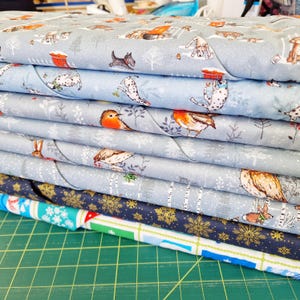 May include: A pile of patterned fabric with winter-themed designs. The fabrics show illustrations of dogs, birds, rabbits, and snowflakes on light blue and navy backgrounds. The bottom fabric has a colourful geometric pattern.