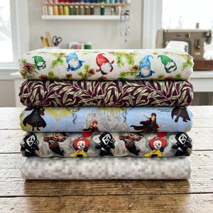 May include: A stack of five folded fabric bolts. The top bolt features gnomes and pine branches. The second has a dark purple leaf pattern. The third displays a light blue wizard school theme. The fourth shows horror movie characters. The bottom is gray pixelated.