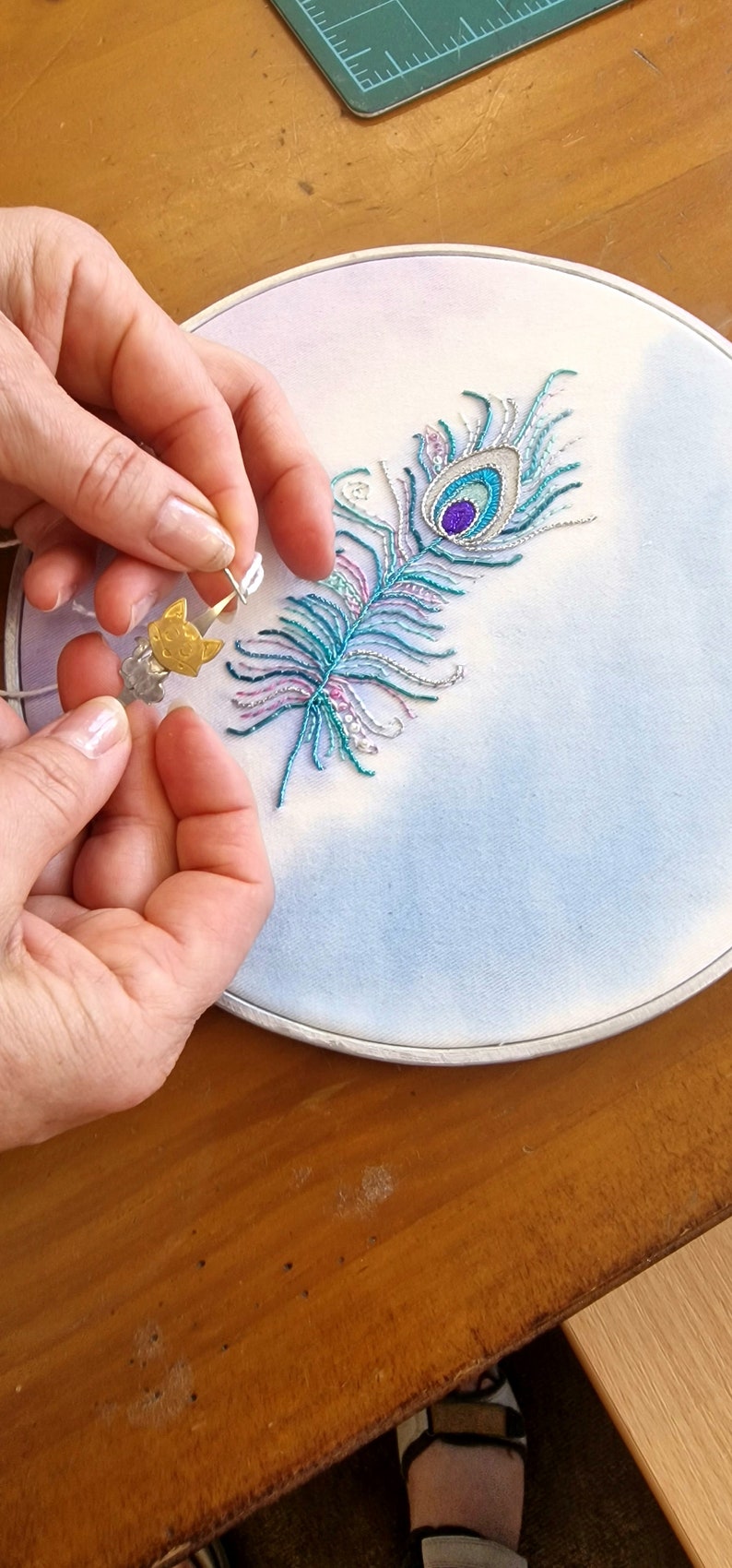 May include: An embroidered peacock feather design in a hoop. The feather is teal, blue, and purple, with silver and pink accents. A small gold cat charm is held near the embroidery.