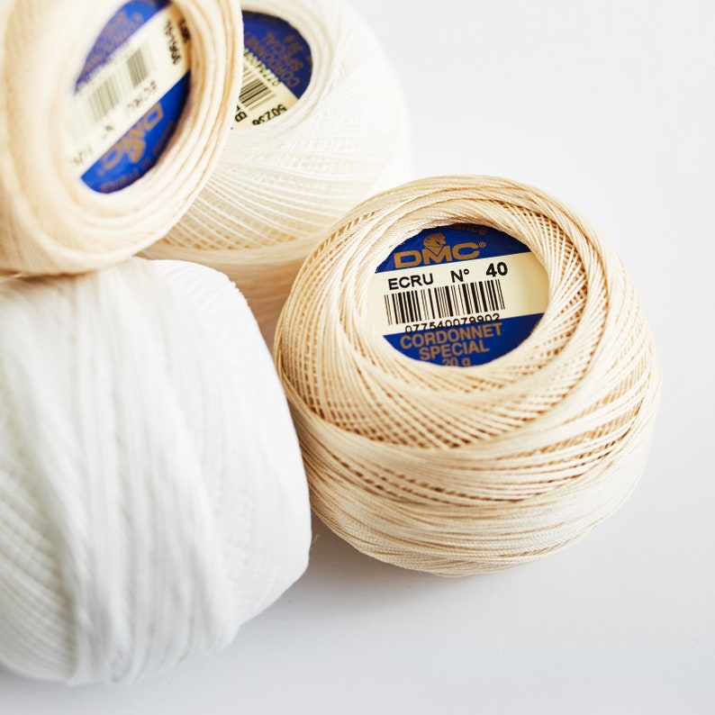 May include: Three spools of DMC Cordonnet Special embroidery floss. One spool is white, and two are a light beige color. The beige spool has a label that reads "DMC, ECRU N&deg; 40, 077540079802, CORDONNET SPECIAL, 20 g."