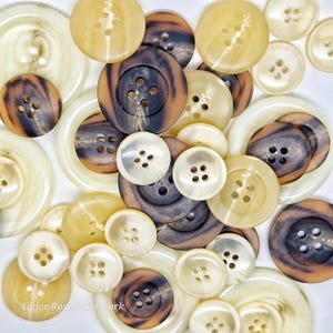 以下が含まれることがあります： クリーム、タン、ブラウンの色合いの、さまざまなサイズのボタンのコレクション。 大理石模様のものもあれば、無地のものもあります。 ボタンには縫い付け用の4つの穴があります。「Tudor Rose Patchwork」の文字が見えます。