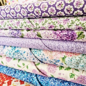 May include: A stack of six fabric swatches featuring floral patterns in shades of purple, blue, and white. The fabrics are arranged in a layered display, showcasing different floral designs and textures.