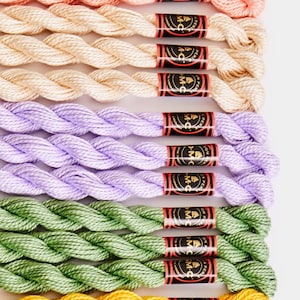 May include: A collection of embroidery floss spools in various colors, including peach, cream, lavender, green, and yellow. Each spool has a black label with the text "DMC" in gold lettering.